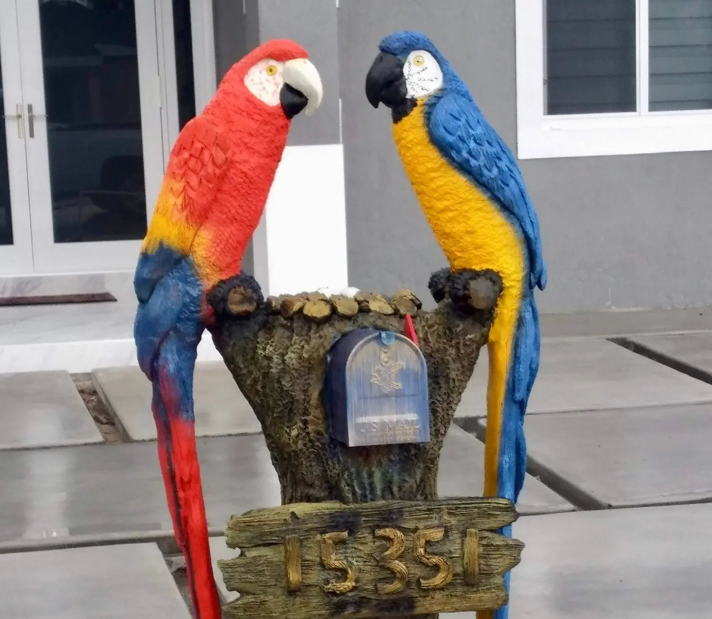 Two colorful parrot statues on a wooden post with a mailbox, set against a neutral background.